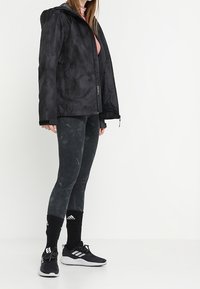 Ung kvinde iført en sort hættejakke, mørkegrå leggings, sorte Adidas-sokker og sorte Adidas-sneakers, der står foran en hvid baggrund.