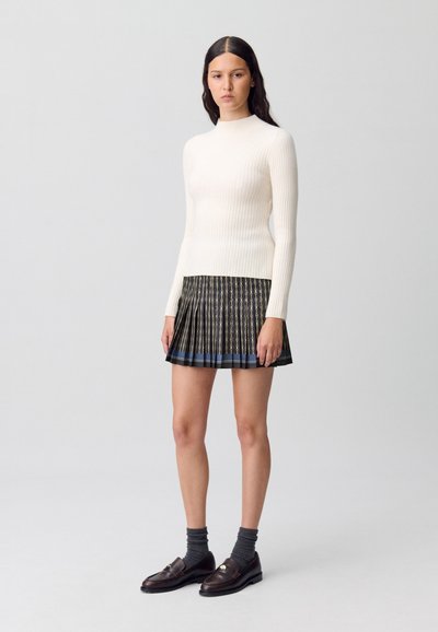 Pull col roulé blanc à côtes associé à une jupe mini plissée noire et dorée. Le modèle porte des chaussettes foncées et des mocassins.