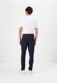 Marineblauwe broek met een gladde textuur en taps toelopende pasvorm, voorzien van een knoopsluiting en twee achterzakken, gedragen met een effen wit t-shirt.