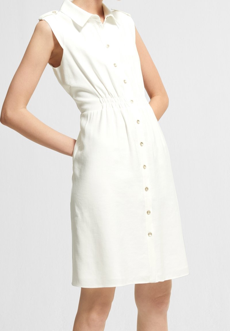 Robe blanche sans manches avec un col, bouton devant et taille élastique. Tissu lisse, légère brillance et boutons en forme de perles.