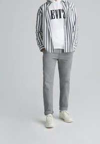 Pasiaste koszula w paski w odcieniach szaro-białych nałożona na białą bluzę z kapturem z logo "LEVI'S", w połączeniu z szarymi spodniami i białymi sneakersami.