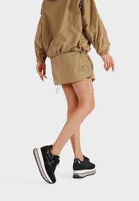 Donna che cammina indossando una gonna e una giacca beige con sneakers a piattaforma bianche e nere su uno sfondo semplice.