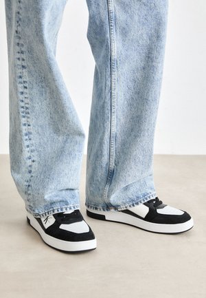 Person iført lyseblå denimjeans med vide ben og sort-hvide sneakers, stående på en beige gulv.