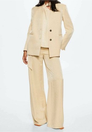 Femme portant un tailleur beige texturé avec un pantalon à jambes larges, un blazer à double boutonnière et un haut assorti avec un col noué.