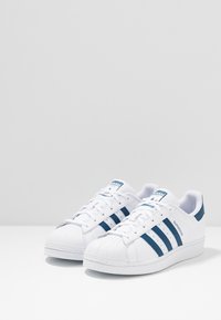 White sneakers with a shell toe, featuring three blue stripes on the sides, a blue heel accent, and a textured sole.