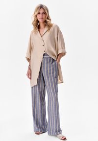 Beżowa, oversize'owa koszula z zapięciem na guziki, połączona z niebieskimi i białymi spodniami o szerokich nogawkach w paski. Gładki materiał i luźny krój.
