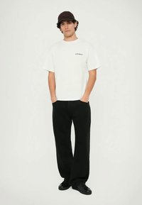 White cotton t-shirt with "No Problemo" text, paired with black wide-leg pants, and a dark brown cap, on a neutral background.