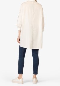 Camicia a bottoni in tessuto leggero beige, con maniche lunghe e taglio morbido, abbinata a jeans skinny blu scuro e scarpe da ginnastica bianche.