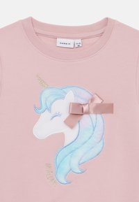 Felpa rosa chiaro con design di un unicorno caratterizzato da una criniera pastello, corno dorato e un dettaglio a fiocco di raso di lato. Il testo recita "unicorno."