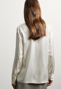 Blusa di seta bianca a maniche lunghe con colletto standard, maniche con polsini e una texture liscia, caratterizzata da un taglio dritto e dettagli di cucitura sul retro.