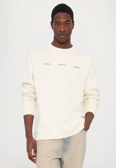 Kreemi värvi crewneck-sviiter musta tekstiga "HZW (25-26) HOLZWEILER "MÄLU." Pehme tekstuur ja lõdva lõikega, ribidega mansetid.