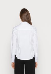 Biała koszula z długim rękawem i kołnierzykiem, o gładkiej fakturze i prostym kroju, wykończona mankietami na guziki oraz hemem o różnej długości.