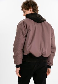 Överdimensionerad mauve bomberjacka med svart huva, ribbade muddar och en texturerad foder. Innehåller sidofickor och en rundad fåll.