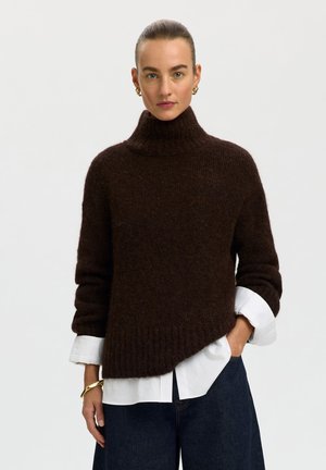 Donna che indossa un maglione dolcevita marrone scuro sopra una camicia bianca, abbinato a pantaloni blu scuro, orecchini a cerchio dorati e un braccialetto.