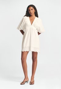 Weißes Kleid mit Puffärmel, V-Ausschnitt, strukturiertem Stoff und gewelltem Saum. Verfügt über gestickte Details überall. Kombiniert mit schwarzen Sandalen.