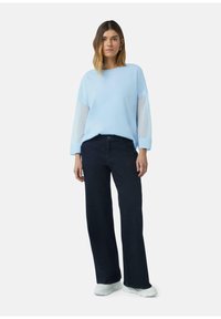 Lichtblauwe trui met doorzichtige lange mouwen, gecombineerd met donkere blauwe wijde jeans en witte sneakers. Effen achtergrond.