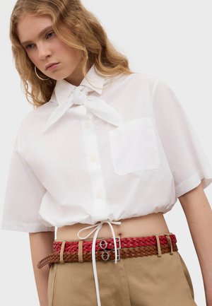 Young person with wavy blonde hair wears a cropped white button-up shirt with front tie, paired with beige pants and two braided leather belts.
