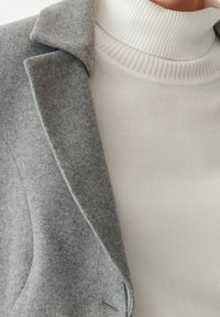 Close-up van een witte geribde coltrui gecombineerd met de lapel van een grijze wollen jas, waarbij de stofstructuur en het lagen effect worden benadrukt.