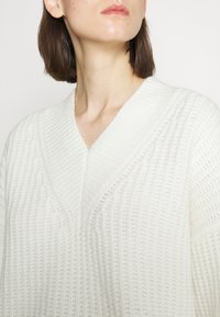 Pull en tricot blanc avec un décolleté en V profond, présentant un motif texturé et une finition côtelée. Tissu doux avec une apparence confortable.