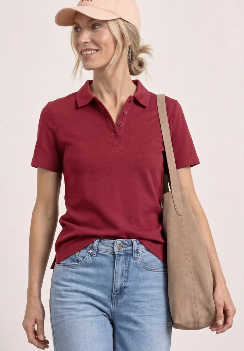 Femme portant un polo rouge à manches courtes, un jean bleu clair, une casquette beige et portant un grand sac cabas beige sur l’épaule, souriant à gauche.