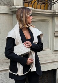 Schwarzes Blazer mit weiten Ärmeln, enganliegendes schwarzes Oberteil, weißer Schal über den Schultern drapiert und eine cremefarbene, gesteppte Handtasche mit goldenen Akzenten.