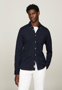 Camicia a maniche lunghe blu navy con colletto a bottoni, pattina frontale, bottoni bianchi e dettaglio con logo. Tessuto liscio abbinato a una maglietta bianca sotto.