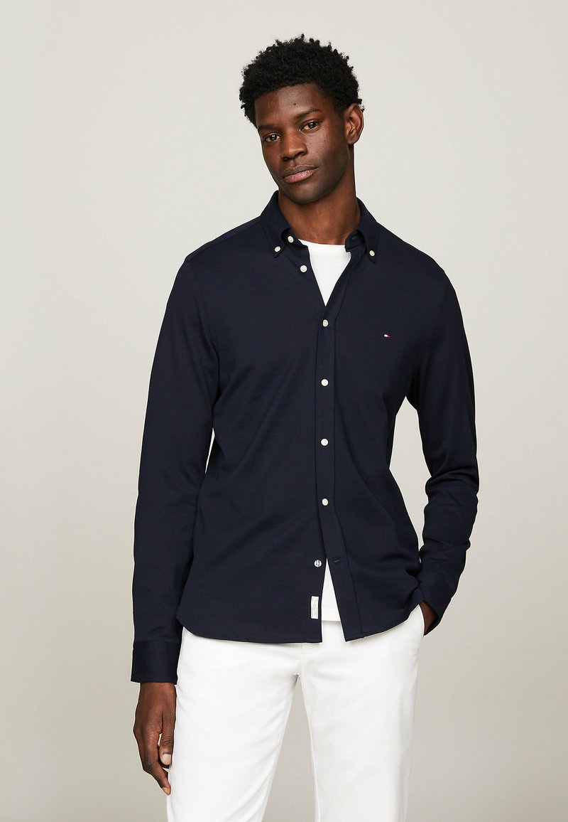 Camicia a maniche lunghe blu navy con colletto a bottoni, pattina frontale, bottoni bianchi e dettaglio con logo. Tessuto liscio abbinato a una maglietta bianca sotto.