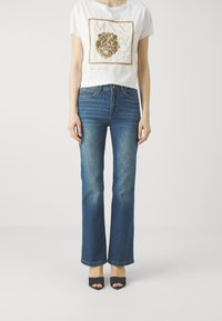 Camiseta blanca corta con gráfico de león dorado en el frente, combinada con jeans acampanados azules y tacones negros destalonados. Textura de denim suave.