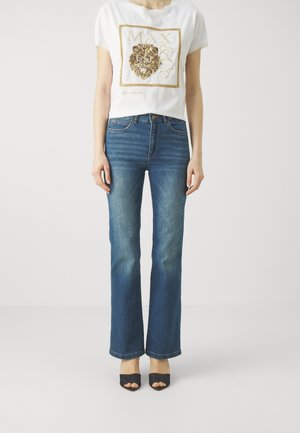 White cropped t-shirt with gold lion graphic on front, paired with blue flared jeans and black open-toe heels. Smooth denim texture.