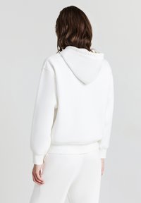 Person mit mittelbraunem Haar, der einen schlichten weißen Hoodie und weiße Hosen trägt, steht mit dem Rücken zu einer schlichten hellen Hintergrund.