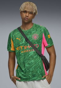 Grön kortärmad tröja med abstrakta mönster, med orange, rosa och marinblå accenter. Visar "ETIHAD AIRWAYS" i rosa bokstäver.