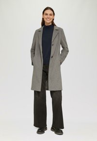 Grau melierter Wollmantel mit Reverskragen, doppelter Knopfleiste und seitlichen Taschen, getragen über einem marineblauen Pullover und schwarzen weit geschnittenen Hosen.
