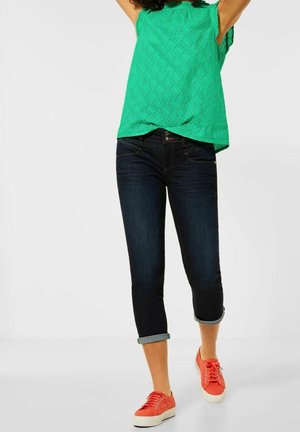 Haut en dentelle verte à manches courtes, jean bleu foncé retroussé aux chevilles et baskets orange. Accent sur les contrastes de texture et de couleur dans la tenue.