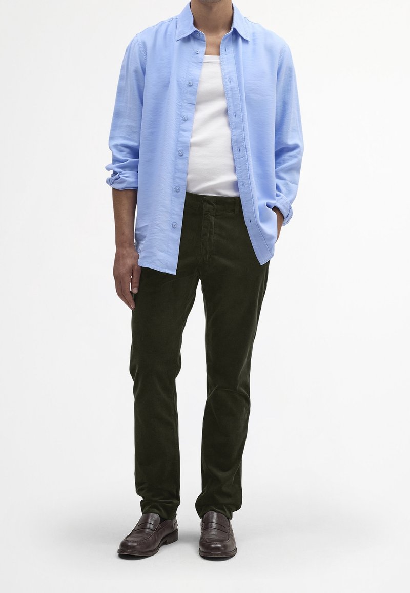 Homme portant une chemise bleu clair à boutons sur un débardeur blanc, un pantalon vert foncé et des mocassins en cuir marron, debout avec une main dans la poche.