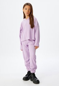 Chica vestida con un hoodie de punto lila y pantalones a juego, con botas de encaje negras, de pie contra un fondo blanco liso.