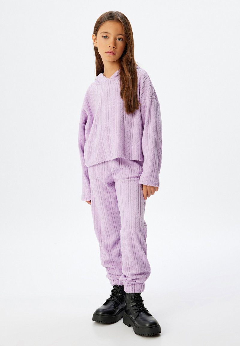 Chica vestida con un hoodie de punto lila y pantalones a juego, con botas de encaje negras, de pie contra un fondo blanco liso.