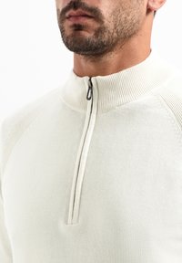 Lichtbeige ritsvest met een hoge geribde kraag. Beschikt over een gedeeltelijke voorrits en een gladde textuur met subtiele naaddetails.