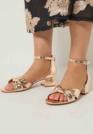 Voeten met gouden metallic enkellaarsjes met blokhak en gedraaide voorste bandjes en een zwarte rok met beige bloemenborduursel.
