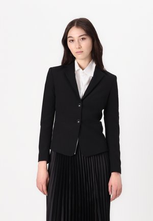 Young woman in a black blazer and white shirt stands against a plain background, looking directly at the camera, hands relaxed by her side.