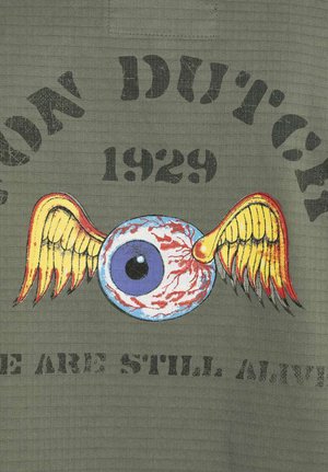 Un design graphique sur un tissu vert olive présente un œil stylisé avec des ailes en rouge, jaune et bleu, accompagné d'un texte noir en gras.