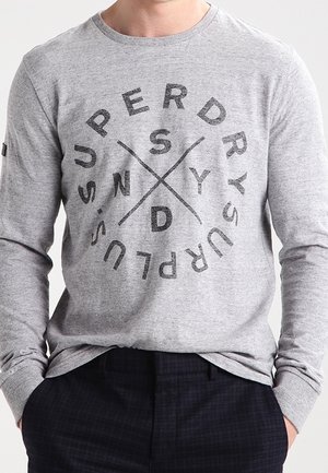 Osoba w szarej, długorękawowej bluzie z okrągłym czarnym napisem "Superdry" z przodu, ręce w kieszeniach ciemnych spodni.