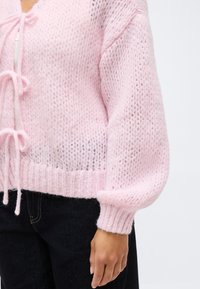 Ροζ μαλακό πλεκτό ζακέτα με μακριά μανίκια και δεμένους κλεισίματα μπροστά, φορεμένη με σκούρα τζιν μπροστά σε απλό φόντο.