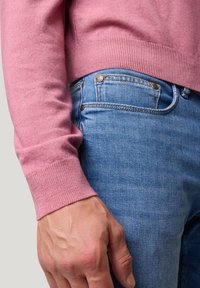 Rosafarbener gestrickter Pullover mit gerippten Bündchen, kombiniert mit hellblauen Jeans. Eine Nahaufnahme zeigt eine Hand, die auf den Jeans ruht und die Textur sowie die Nähdetails sichtbar macht.