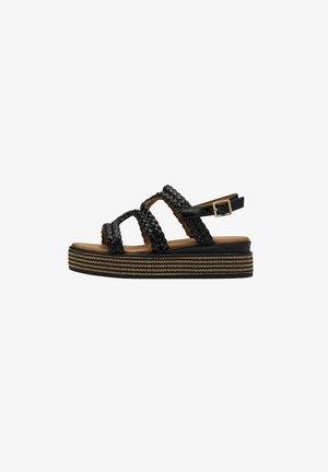 Sandales avec sangle tressée noire et semelle plateforme, présentées avec une bande accent texturée dorée. Sangle de cheville ajustable avec une boucle en métal.