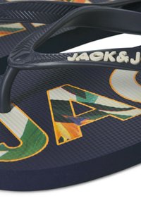 Primo piano di un cinturino di ciabatta nera su una suola testurizzata con le lettere colorate "JACK & J" parzialmente visibili sulla superficie della suola.