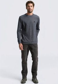 Pánska tmavosivá dlhorukávová košeľa s voľným strihom, okrúhlym výstrihom a malým logom na hrudi, v kombinácii s tmavými džínsami a zelenými topánkami.