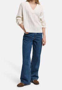 Pull en tricot crème à col en V avec texture côtelée, associé à un jean denim bleu à jambes larges. Chaussures décontractées avec des accents marron et orange.