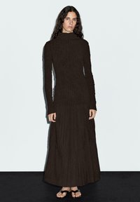 Black long-sleeve dress with a fitted top and loose pleated skirt. Textured fabric with a subtle pattern, paired with flat black sandals.