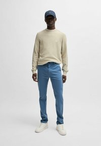 Beige textured sweater, blue pants, and white sneakers. Completing the look is a navy cap with a logo patch.