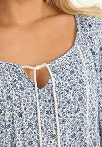 Kukka-aiheinen paita valkoisena ja sinisenä, jossa on V-pääntie nauhatyylillä ja kerrostettu rakenne. Kangas näyttää kevyeltä ja hengittävältä.